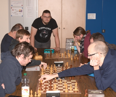 Rechts und links in Reihe einander gegenüberssitzend je drei Schachspieler.
    Zwischen ihnen der Tische mit den Schachpartien. Am Ende der Tischreihe
    ein Kibitz.