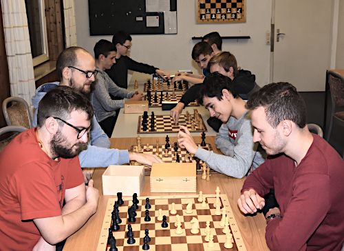 An beiden Seiten der Tischreihe sitzen jeweils 5 Schachspieler
        einander gegenüber und spielen ihre Partien.